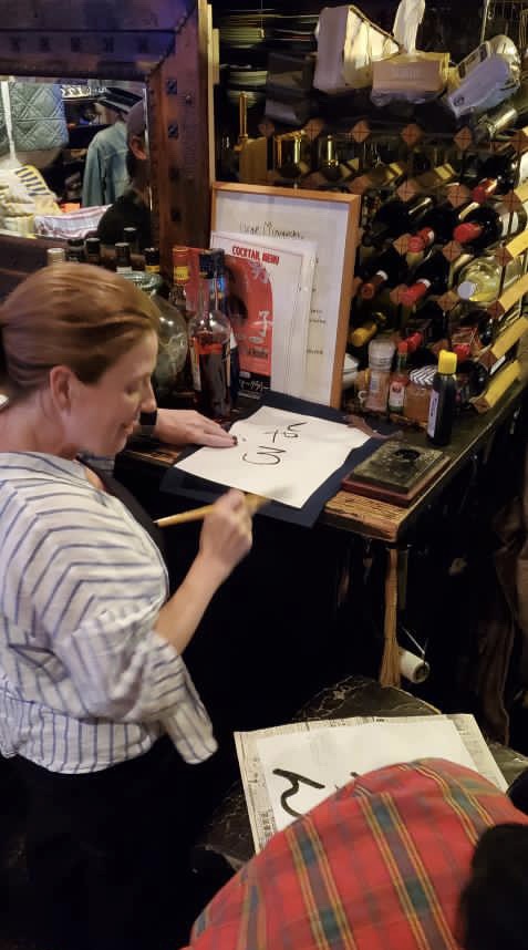 A woman wearing a striped shirt is writing on paper with a brush in a dimly lit environment, surrounded by shelves of bottles and other items.