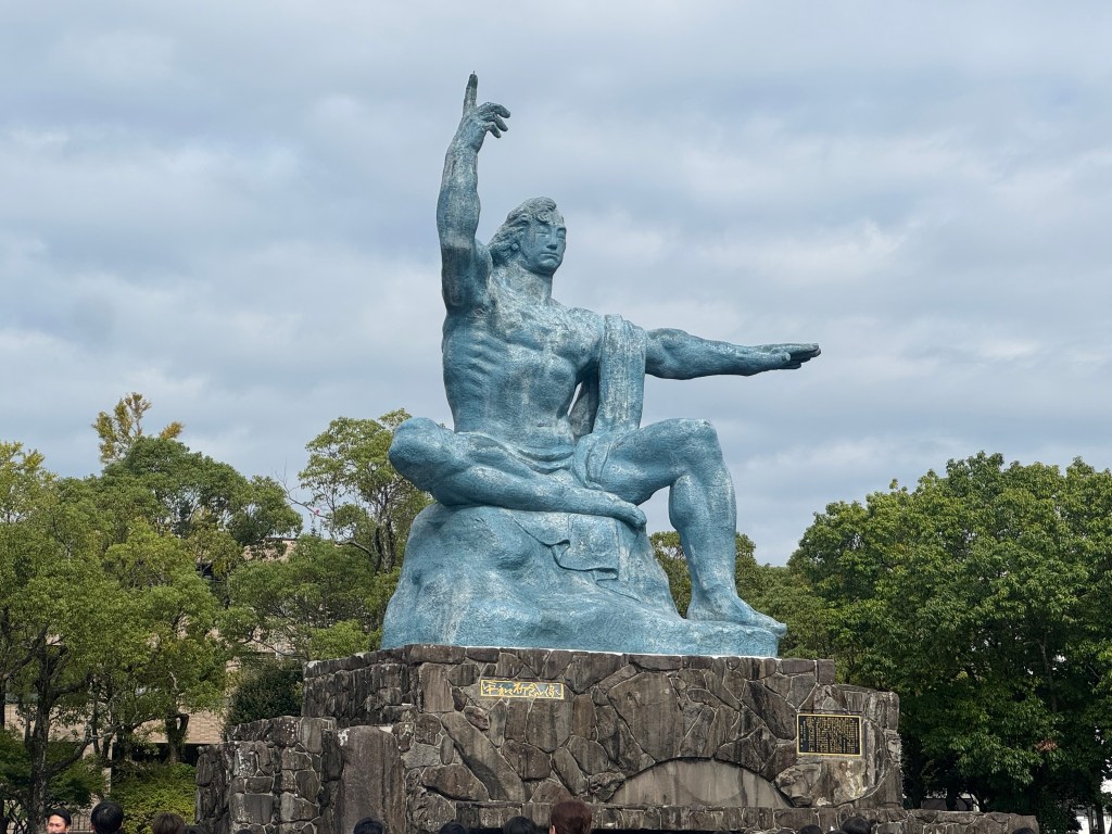 A large bronze statue of a seated figure with one arm raised, surrounded by green trees and a cloudy sky, with visitors gathered around.