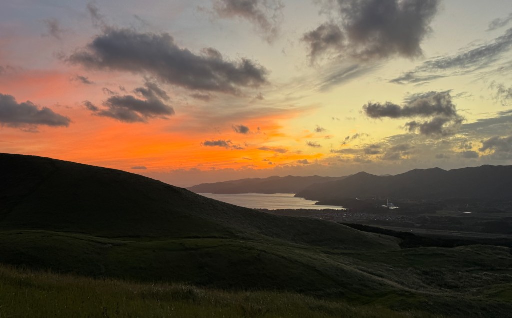 A scenic sunset over rolling hills and a body of water, with vibrant orange and pink hues illuminating the sky.