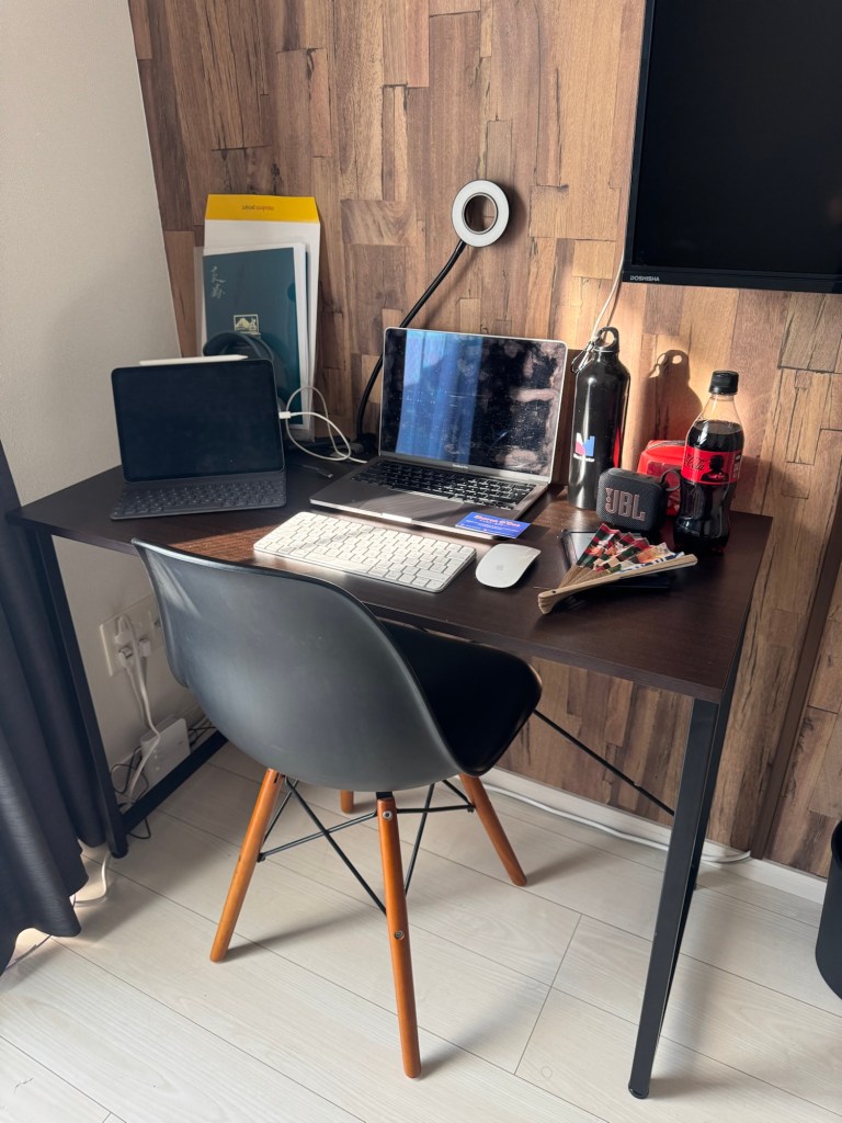 A workspace setup featuring two laptops, a keyboard, a water bottle, and a can of soda on a wooden desk with a wall-mounted light in a well-lit room.