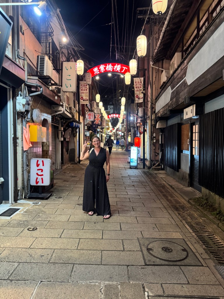 A person smiling and posing in a narrow, dimly lit street lined with traditional lanterns and signs, showcasing a lively nighttime atmosphere in Japan.