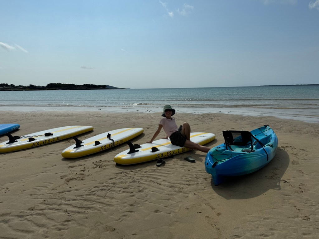 A person sitting on the beach next to paddleboards and a kayak, with a calm sea and blue sky in the background.