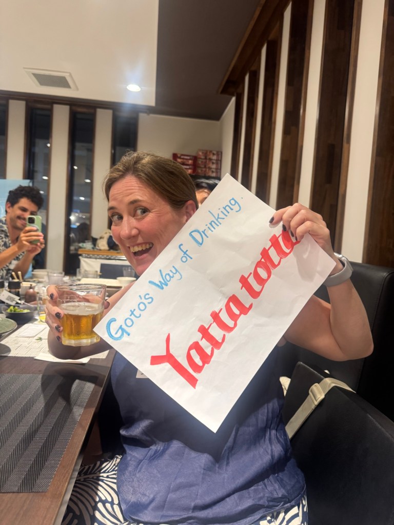 A person smiling while holding a sign that says 'Goto's Way of Drinking Yattatottu' and a glass of beer, in a restaurant setting.