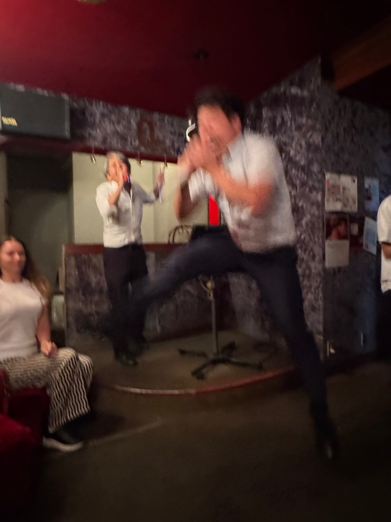 A group of people having fun at a karaoke bar, with one person energetically jumping on stage while another sings into a microphone.