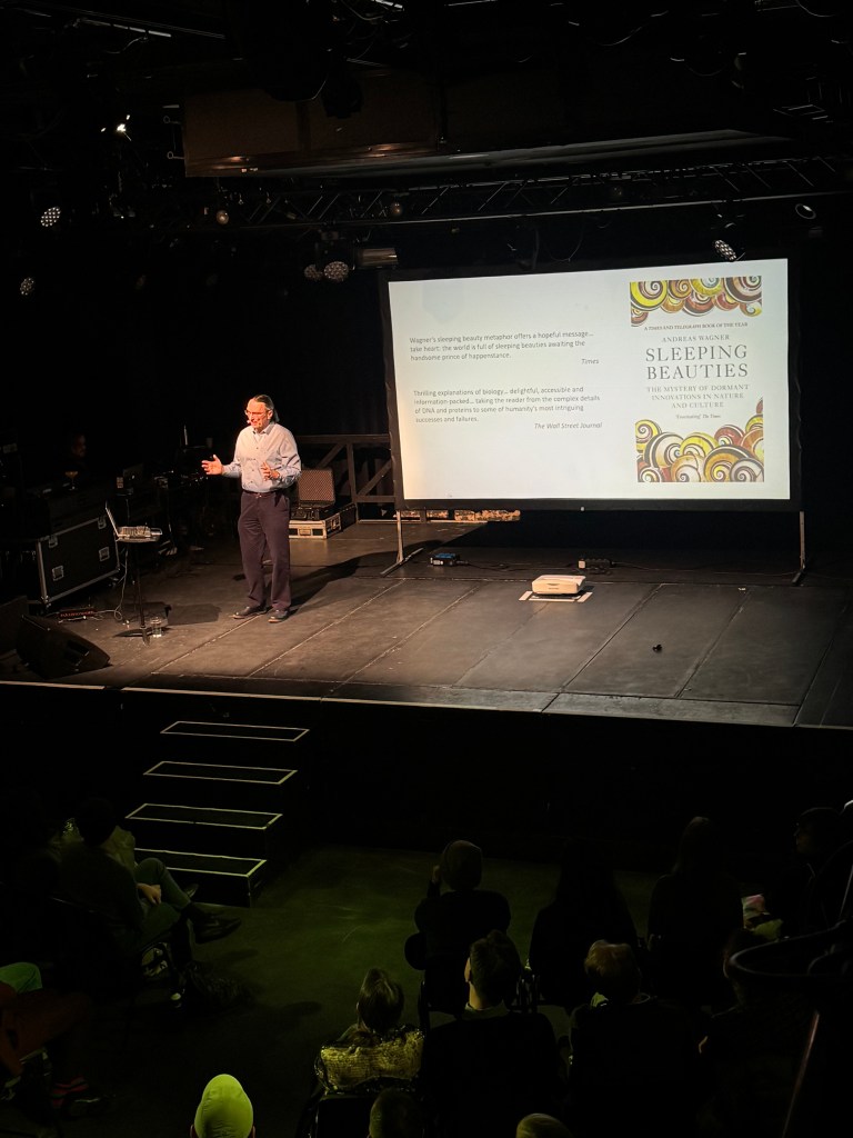 A speaker presenting on stage at an event, discussing the topic of dormant innovations. A presentation slide is visible in the background with the title 'Sleeping Beauties'. The audience is seated in front of the stage, engaging with the talk.