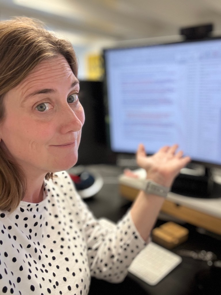 A woman with shoulder-length brown hair and a polka-dotted shirt is sitting at a desk, gesturing with her hands in a questioning manner, while looking at the camera. A computer screen displaying text is visible in the background.