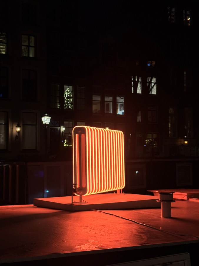 A glowing neon radiator sculpture illuminated against a dark urban backdrop, showcasing the architectural features of nearby buildings.