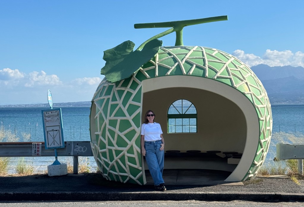 A person stands next to a fruit-shaped bus stop designed to look like a pineapple, with a coastal view in the background.