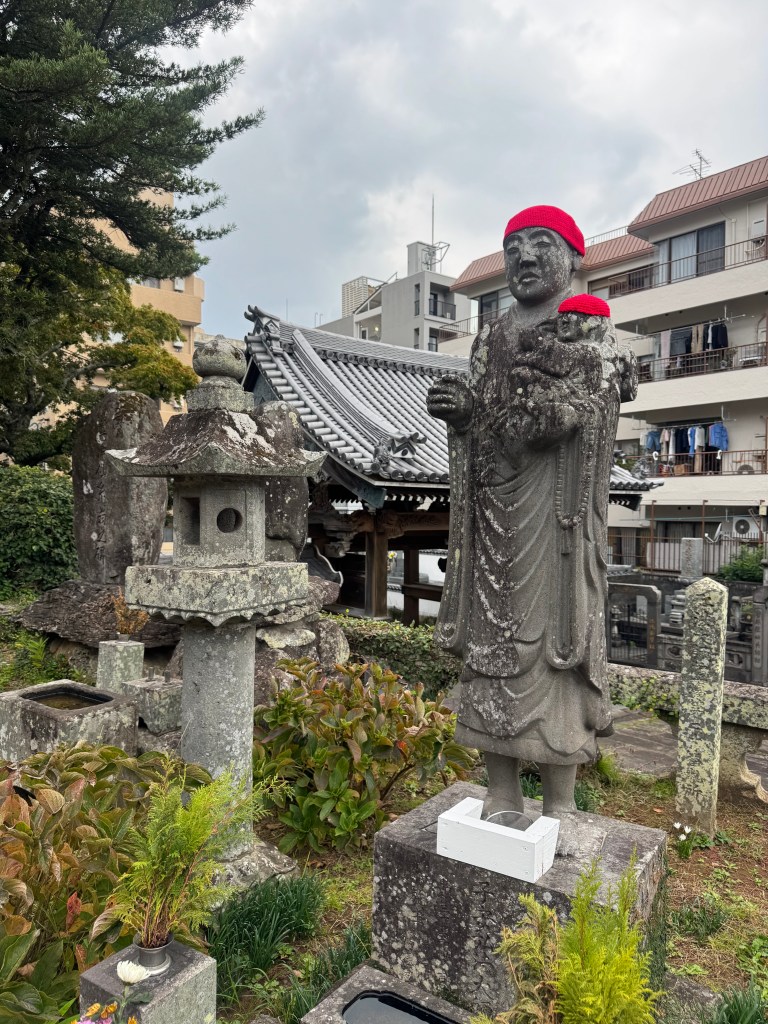 A stone statue of a figure wearing a red hat, surrounded by greenery and traditional Japanese architecture, in Nagasaki, Japan.