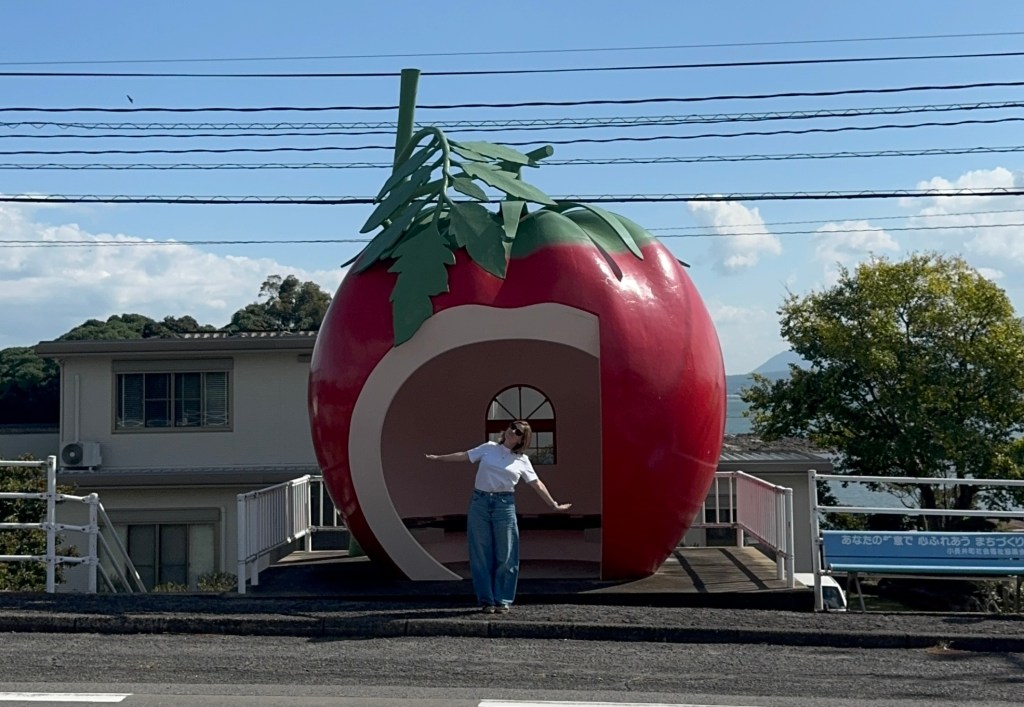 A person standing in front of a large, fruit-shaped bus stop designed like a tomato, with a clear blue sky and greenery in the background.