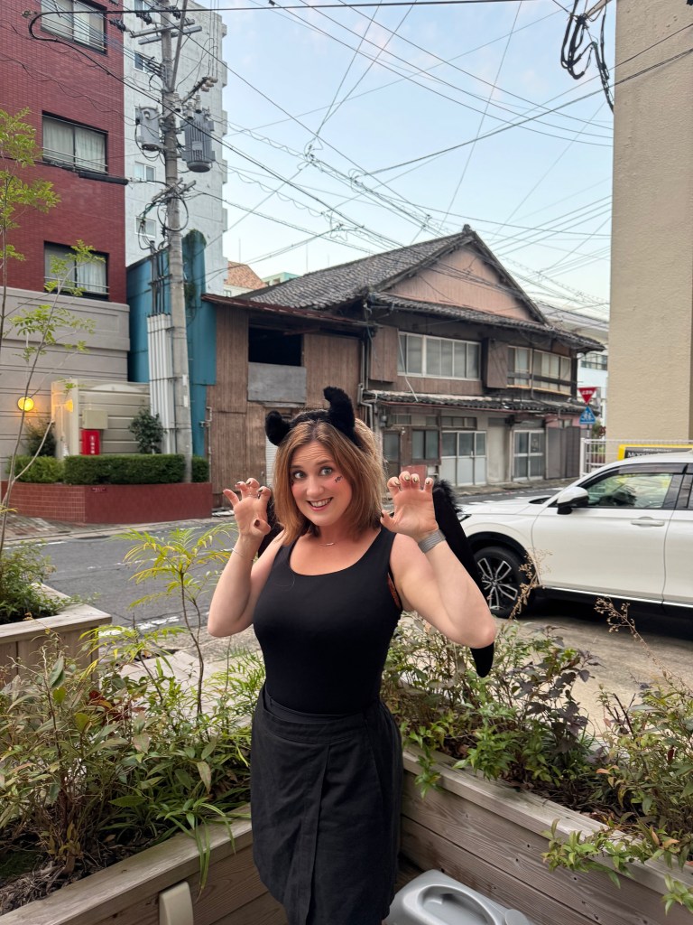 A person dressed in a black outfit and cat ears playfully poses with claw-like hands against a backdrop of urban buildings and electric wires.