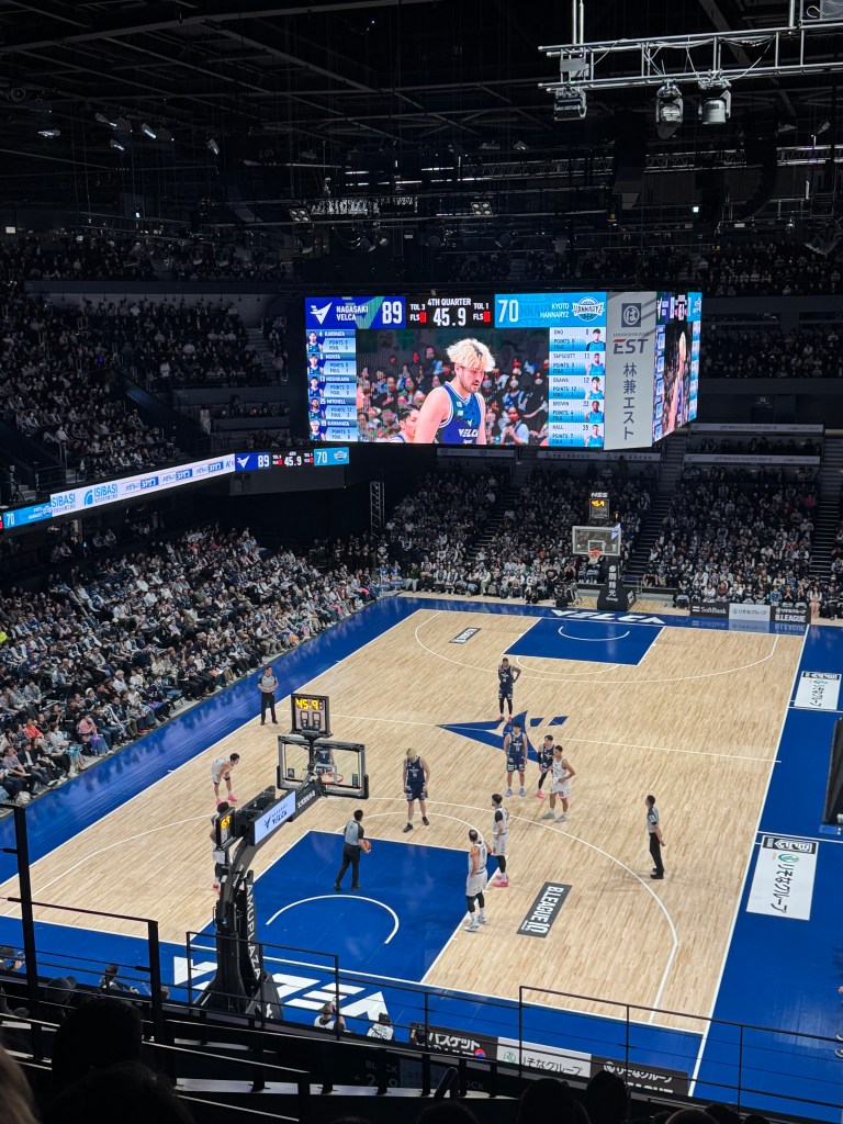 A basketball game in a stadium with a large scoreboard displaying the score, players in action, and a packed audience.