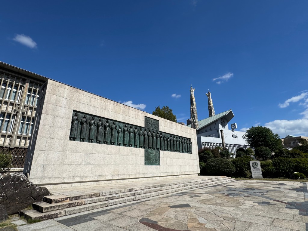 A monument commemorating the 26 martyrs in Nagasaki, with statues of figures on a granite wall against a clear blue sky.