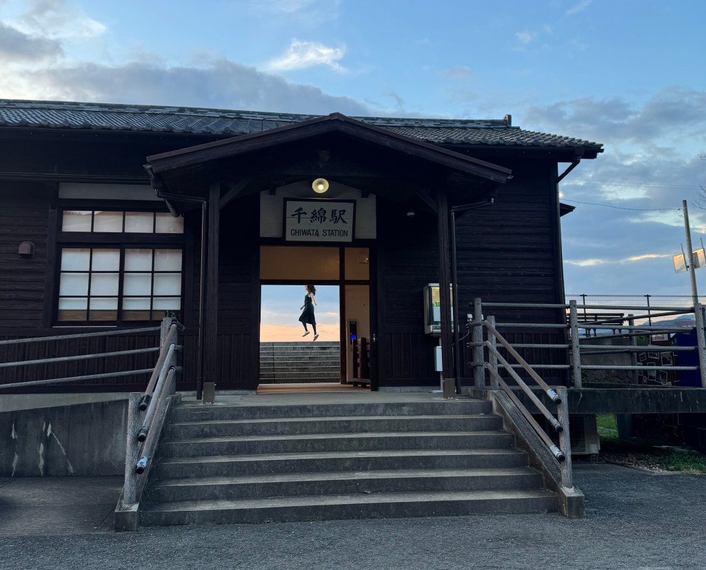 An image of Chiwata Station, a traditional wooden train station with a person walking up the steps, framed by a sunset sky.