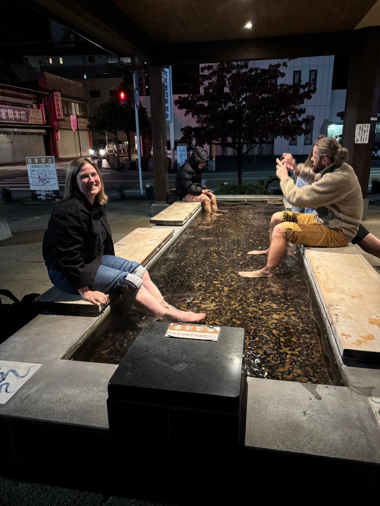 A group of people enjoying foot baths in a public onsen area at night, with one person smiling at the camera.