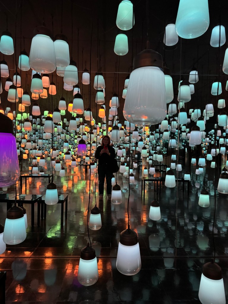 A woman standing in an art installation with colorful hanging lamps, reflecting light on a shiny floor.