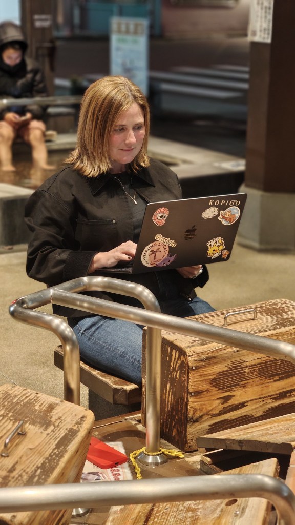 A person sitting at a wooden bench with their laptop, focused on work, while soaking their feet in a hot spring footbath.