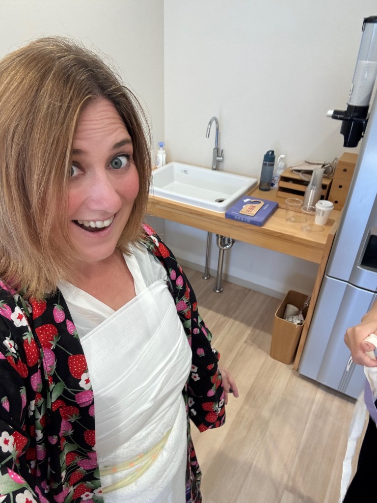 A person smiling while wearing a floral kimono, showcasing a half-dressed traditional outfit in a modern kitchen setting.