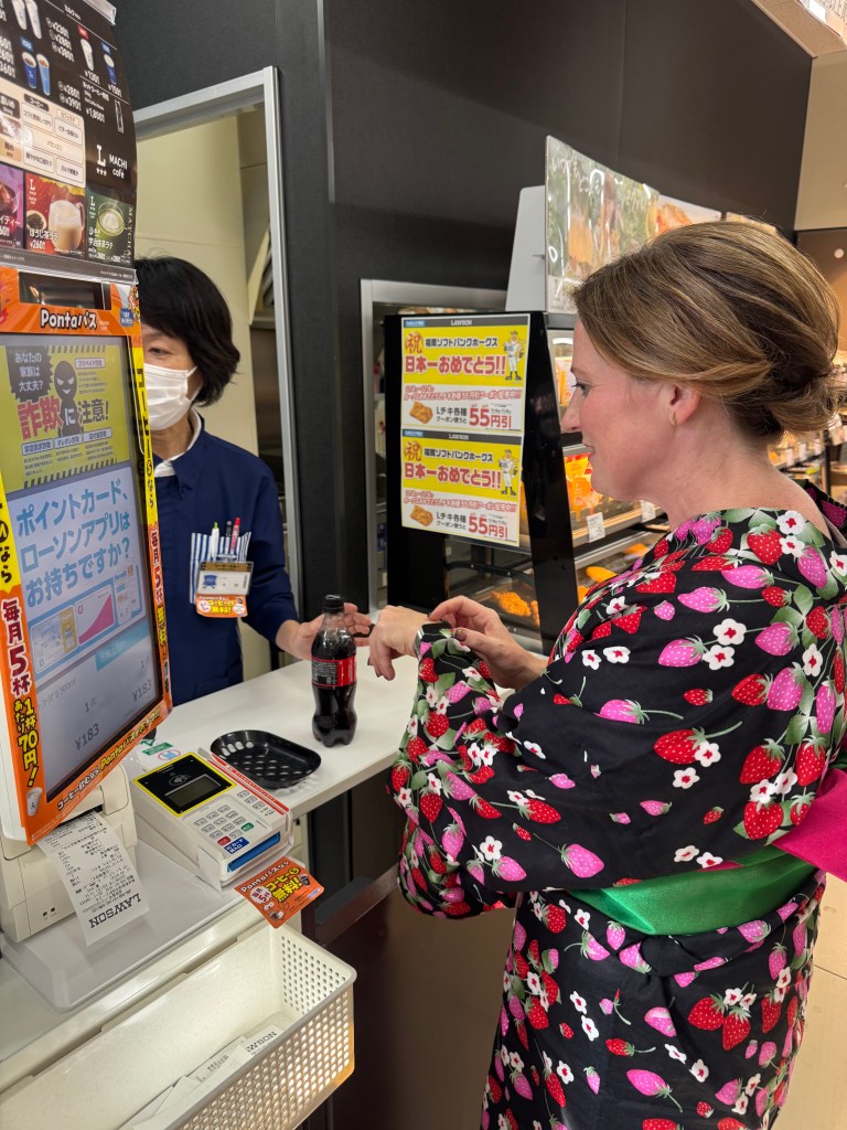 A woman wearing a colorful yukata with strawberry patterns, standing at a convenience store counter, interacting with a cashier while holding a bottle of soda.
