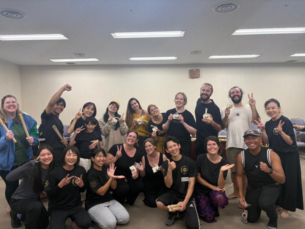 A diverse group of people poses together in a community taiko drumming workshop, holding drumsticks and displaying cheerful expressions.