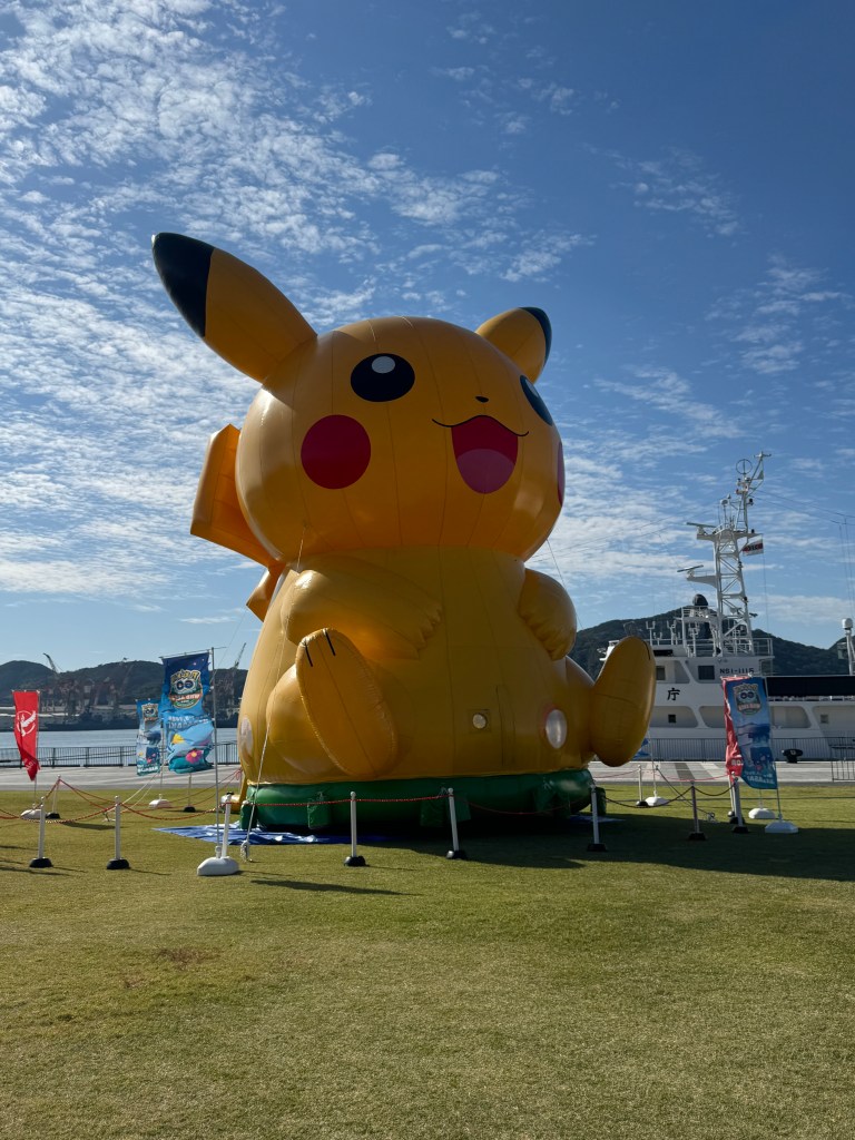 A large inflatable Pikachu standing in a grassy area with a clear blue sky in the background.