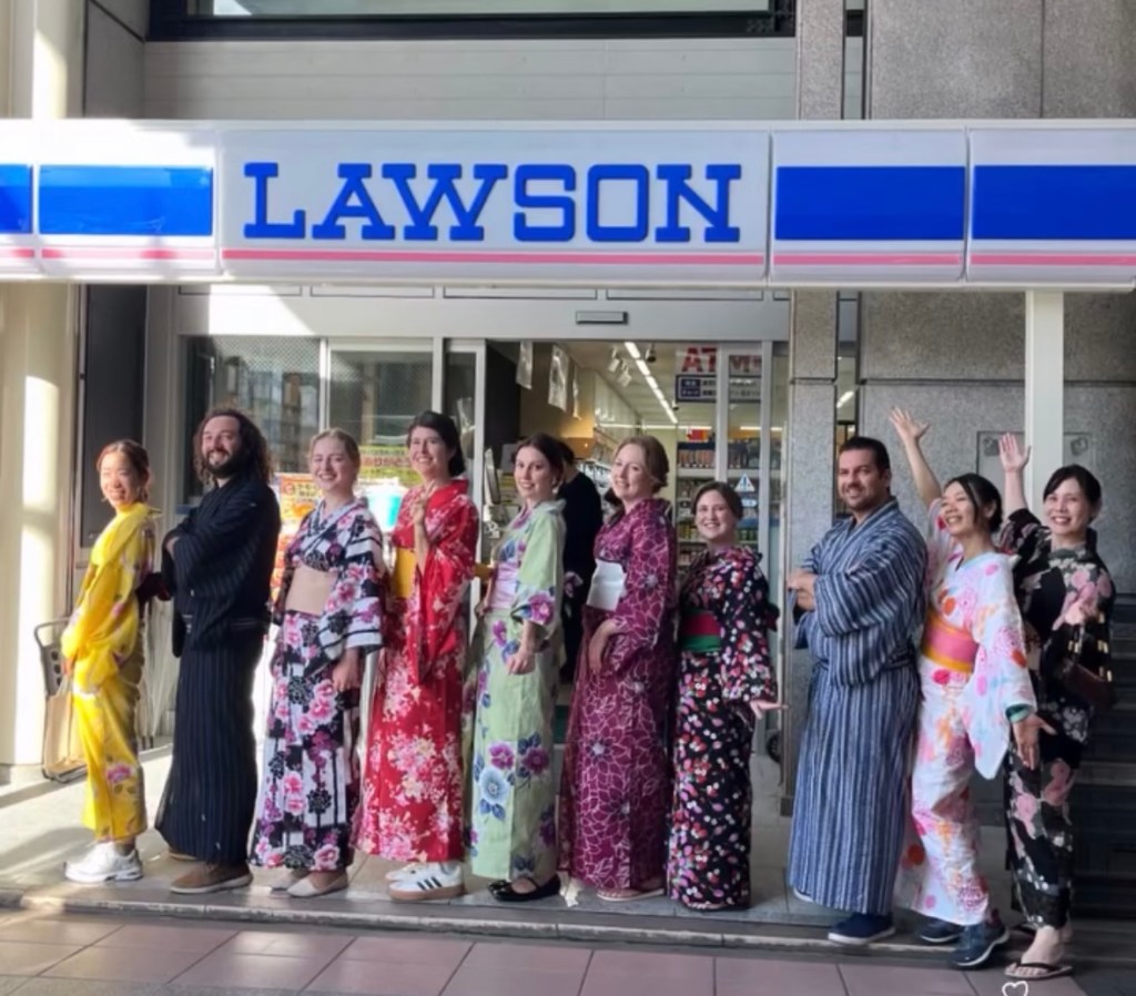 A group of people wearing colorful kimonos stands in a line in front of a Lawson convenience store, smiling and posing for the camera.