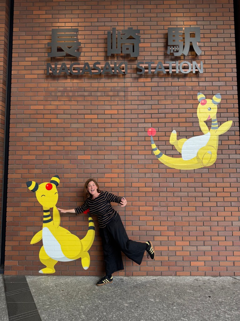A person joyfully posing next to a colorful Pokémon mural at Nagasaki Station, featuring colorful cartoon characters on a brick wall.