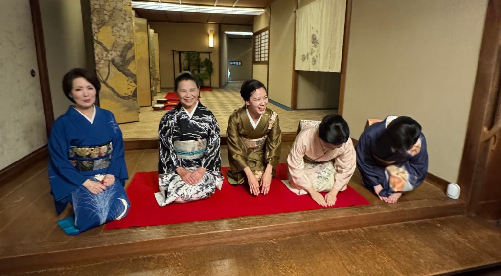 Five women in traditional Japanese kimonos seated on tatami mats, smiling and interacting in a traditional setting.