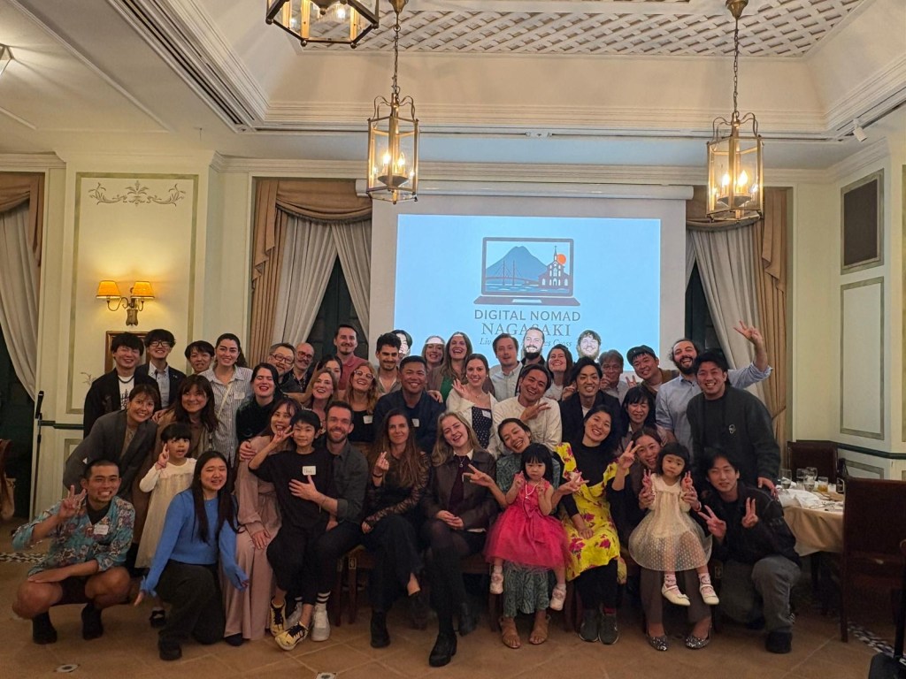 Group photo of participants at the Digital Nomad Nagasaki event, featuring diverse individuals posing together with smiles and a backdrop displaying the event logo.