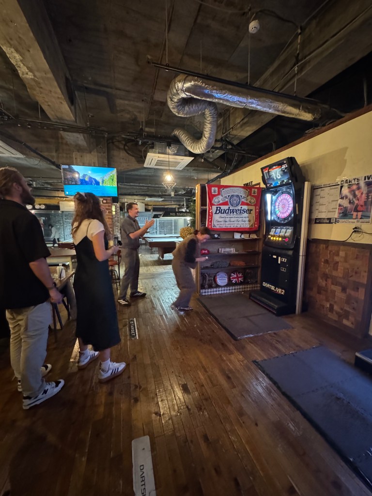 A group of friends enjoying a game of darts in a lively bar setting, with wooden flooring and a Budweiser advertisement visible on the wall.