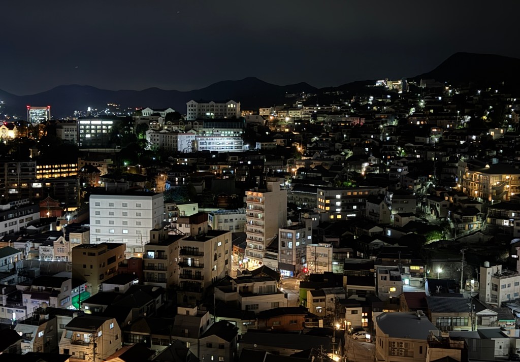A nighttime view of a cityscape, showcasing a dense arrangement of buildings illuminated by various lights, with mountains in the background.