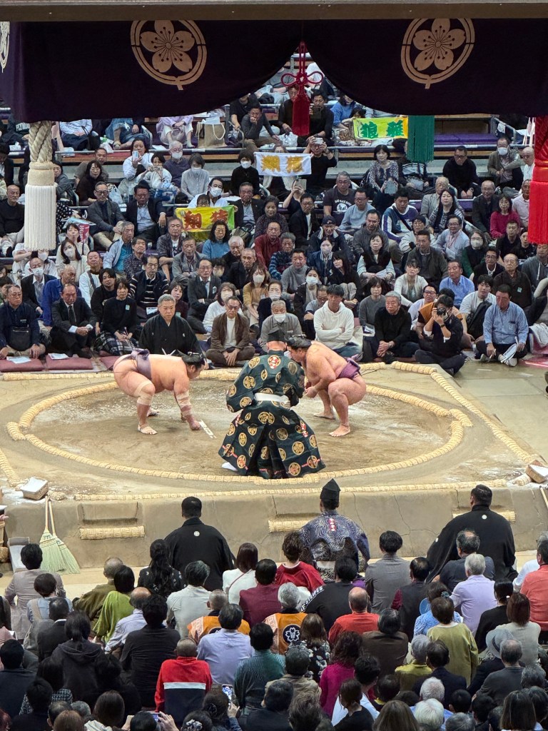 A photo capturing a sumo wrestling match, featuring two wrestlers in the ring amidst a large audience. The wrestlers, wearing traditional mawashi, are in a competitive stance, while a referee in a ceremonial outfit oversees the match. The audience is engaged, with some holding banners. The arena is adorned with traditional decorations.