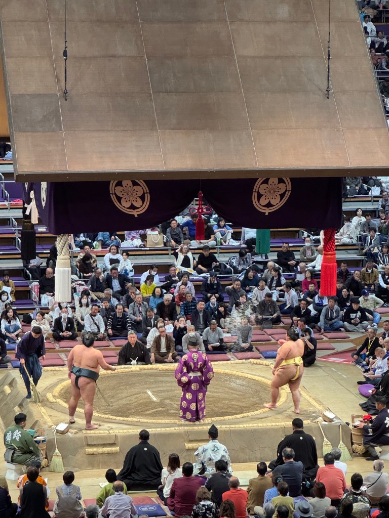 A sumo wrestling match taking place in a traditional arena, with two wrestlers facing off in the ring, surrounded by onlookers. The setting captures the ceremonial atmosphere with spectators seated on tatami mats and a decorated roof above.