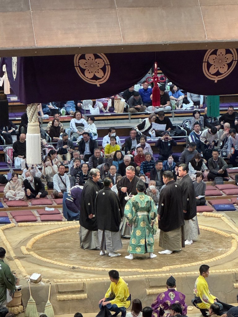 A sumo wrestling ceremony taking place in a circular ring, surrounded by an audience. Participants in traditional Japanese attire are engaged in conversation, while spectators watch eagerly from seated positions.