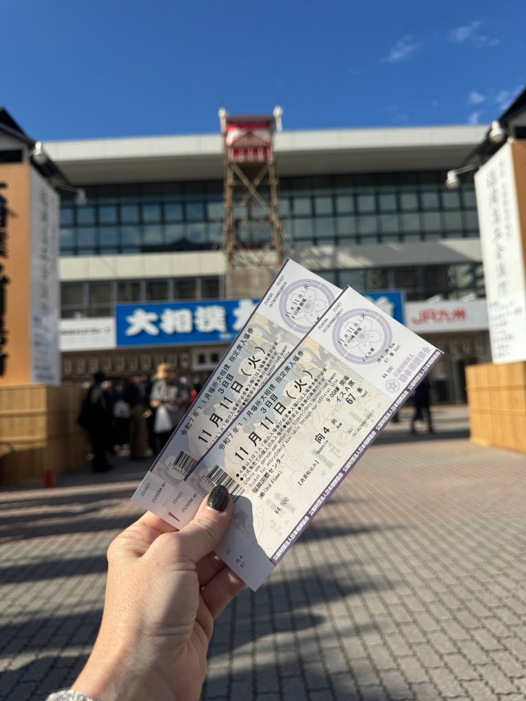 A hand holding two sumo wrestling tickets in front of a venue, with a clear blue sky in the background.