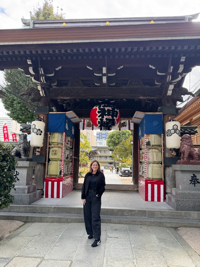 A person standing in front of a traditional Japanese shrine gate, flanked by decorative lanterns and colorful banners, with modern buildings visible in the background.