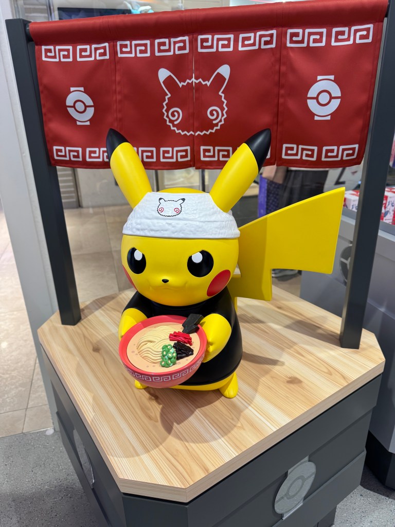 A Pikachu figurine wearing a headband, holding a bowl of ramen on a wooden display, with a red banner in the background featuring Pokémon-themed designs.
