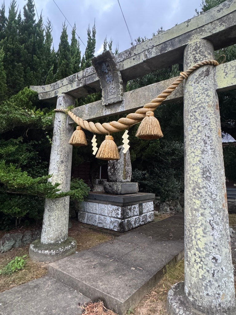 A traditional torii gate adorned with shimenawa rope and tassels, surrounded by dense greenery, symbolizing a threshold between the human world and the spiritual realm.