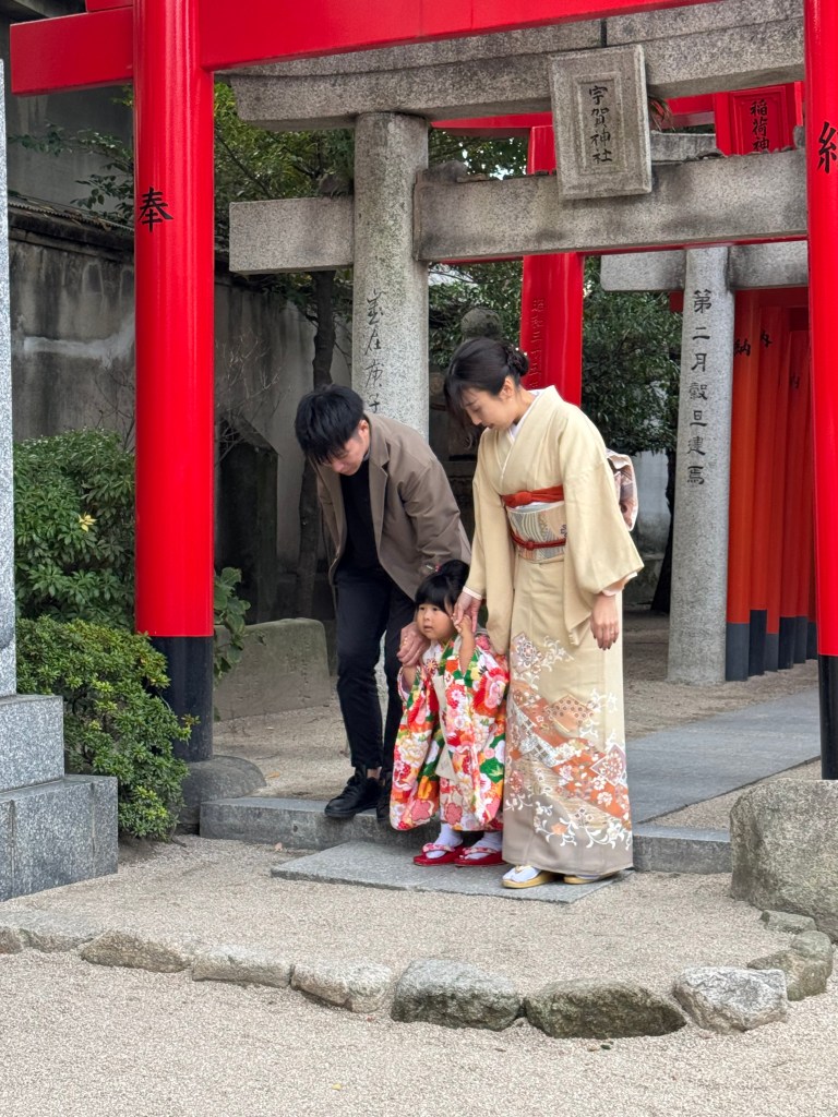 A family at a torii gate in Japan, with a woman in a traditional kimono, a man wearing a casual outfit, and a young girl dressed in a colorful floral garment, all posing together in a serene outdoor setting.