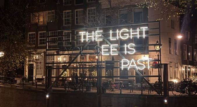 A illuminated sign on a building in Amsterdam that reads 'THE LIGHT YOU SEE IS PAST.' with bicycles parked nearby.