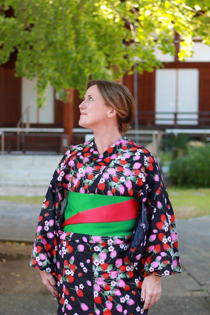 A woman wearing a colorful floral yukata with a green and red obi, looking upwards under a leafy tree.