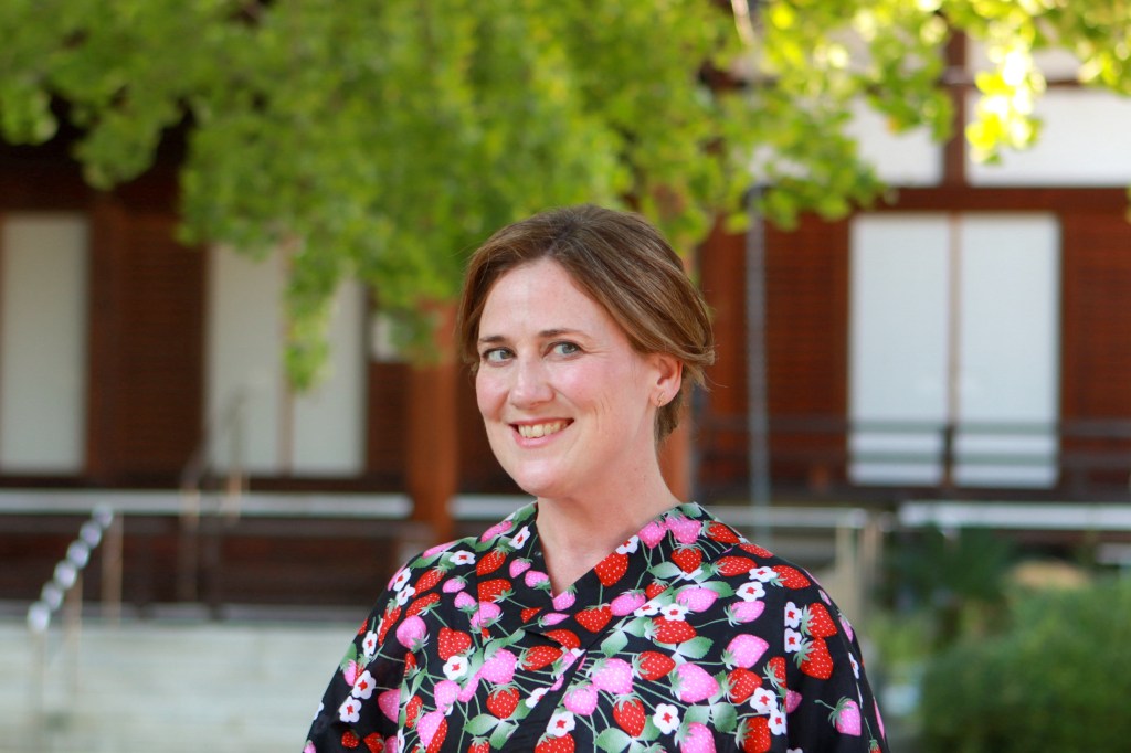 A woman smiling in a colorful floral kimono stands in front of a blurred background of greenery and traditional architecture.