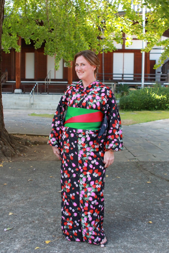 Person wearing a colorful floral yukata with a green obi, standing outdoors under trees.