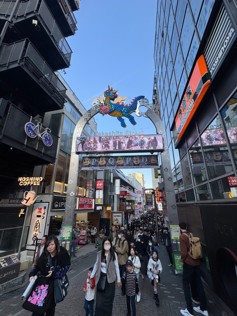 A busy street scene on Takeshita Street in Tokyo, showcasing vibrant shops and a large decorative arch with a dragon design. Crowds of people walk among the stores, creating a lively atmosphere.