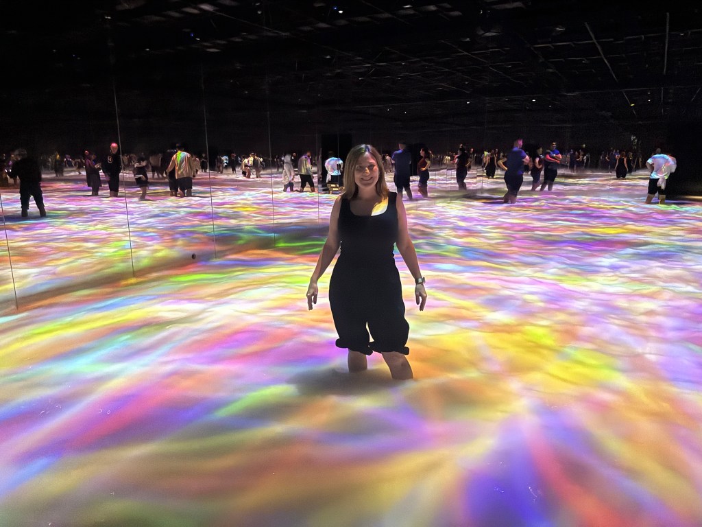 A person standing in a colorful, illuminated room with projections on the floor, surrounded by other visitors.