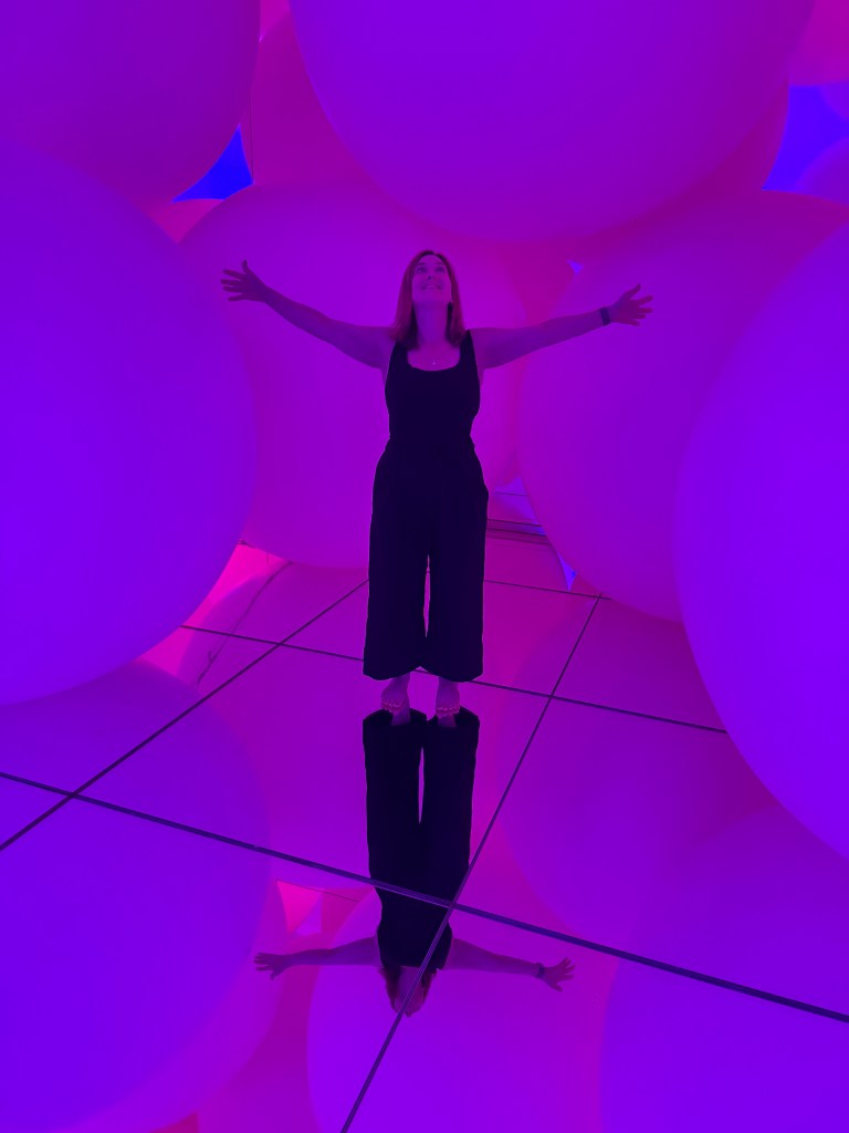 A person stands among large, soft, illuminated spheres, with arms outstretched, in a vibrant purple-toned environment reflecting on a mirrored surface.