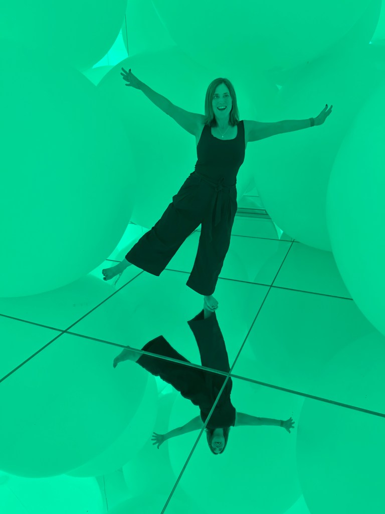 A person standing on a mirrored floor surrounded by large, green spheres, posing with arms spread wide.