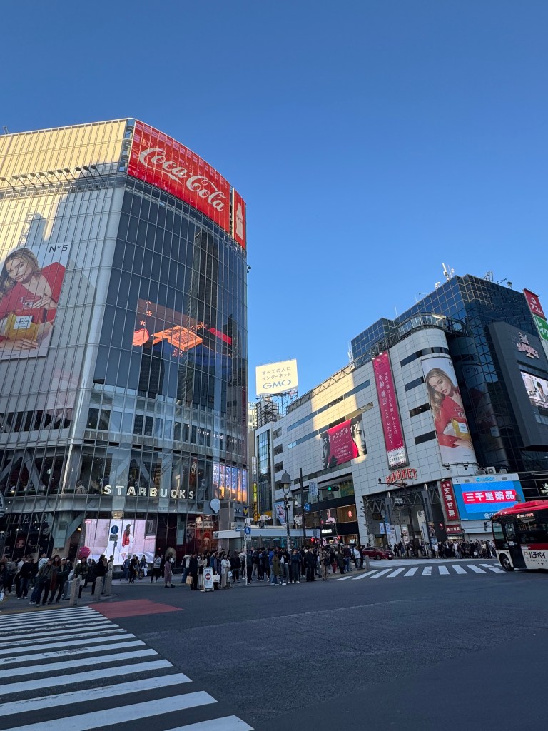 A bustling street scene in Tokyo featuring modern buildings, large advertisement screens, and a crowd waiting at a crosswalk.