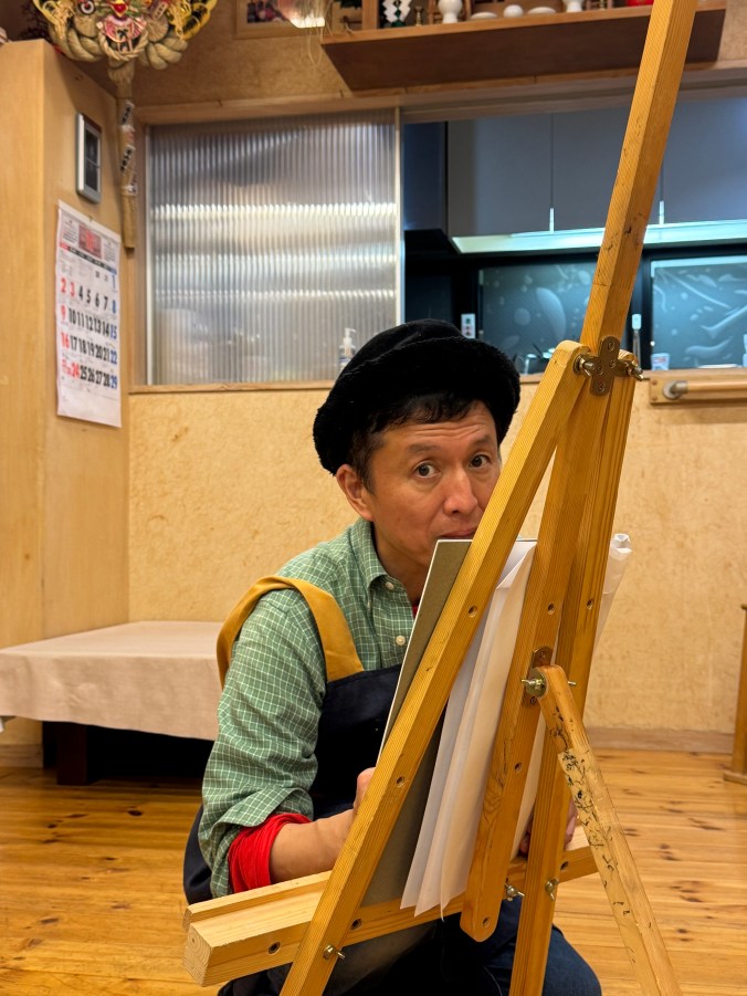 Kagaya sitting at an easel, looking at the viewer with a surprised expression, wearing a green checkered shirt and a black hat, in a cozy, wooden interior.