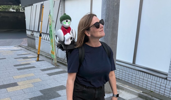 A woman walks down a city street with a small robot on her back, showcasing a blend of technology and urban life.